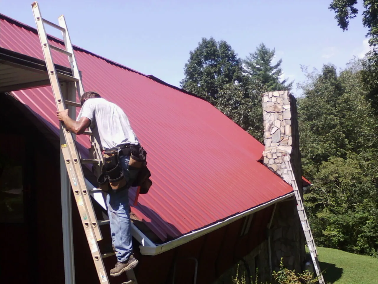 Roofer on a ladder beside a red metal roof and stone chimney — Soffit & Fascia Repair by Roofing Brothers in Ferdinand